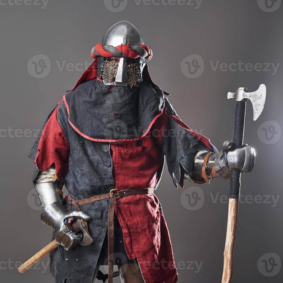 mittelalterlicher Ritter auf grauem Hintergrund. Porträt eines brutalen Kriegers mit schmutzigem Gesicht mit roter und schwarzer Kleidung mit Kettenpanzerrüstung und Streitaxt foto