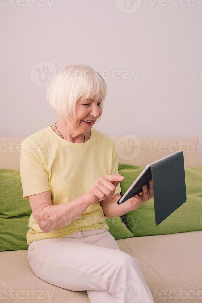 Senior fröhliche kaukasische stilvolle Frau mit grauem Haar mit ihrem Tablet zu Hause. Technologie, Emotionen, Familie, gesunder Lebensstil, positiv denkendes Konzept foto