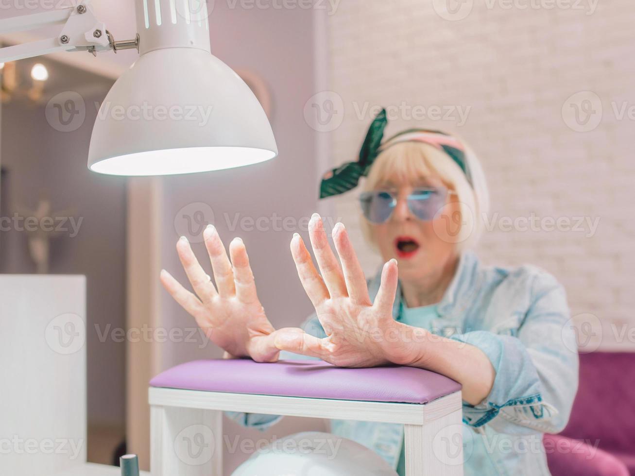 Maniküremeister in blauen Handschuhen, die Hände einer älteren stilvollen Frau in blauer Sonnenbrille und Jeansjacke eincremen, die im Maniküresalon sitzt foto