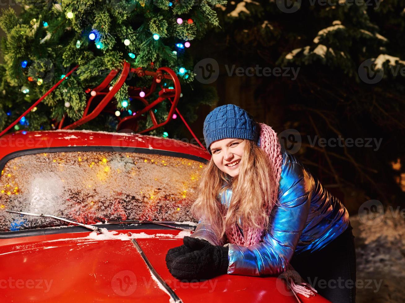 glücklich lächelnde junge kaukasische Frau in Schal, Hut, Jacke, Handschuhe durch den roten Oldtimer und Weihnachtsbaum im Freien. neues Jahr, Spaß, Winterkonzept foto