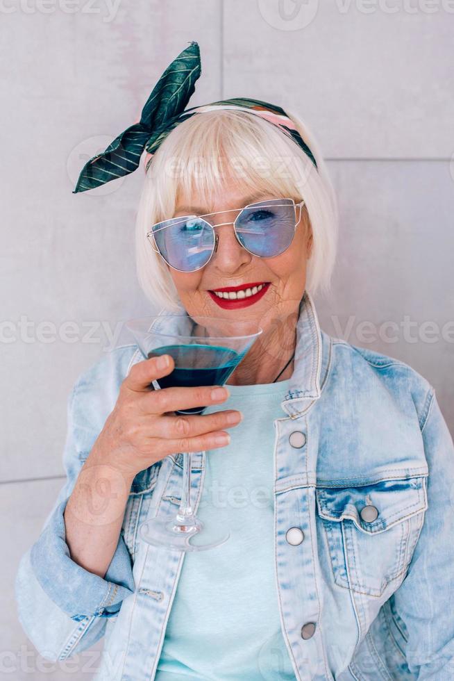 Senior stilvolle Frau mit grauem Haar und in blauer Brille und Jeansjacke mit Glas mit blauem Cocktail. Alkohol, Entspannung, Urlaub, Ruhestandskonzept foto