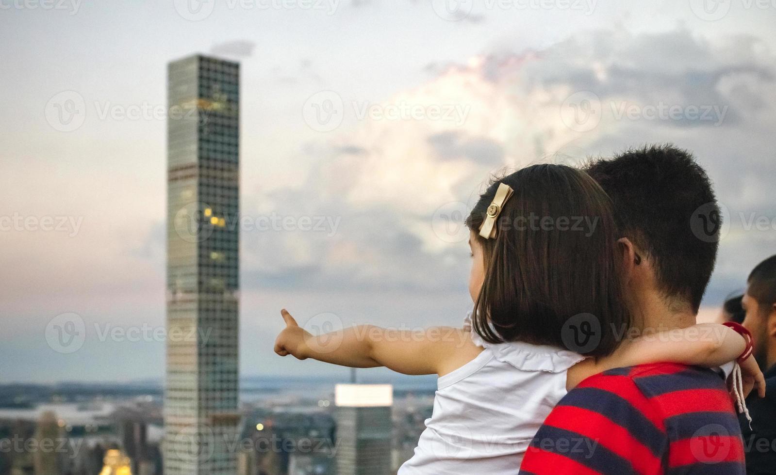 Mman hält kleines Mädchen, das auf den Wolkenkratzer der Skyline von Manhattan zeigt foto