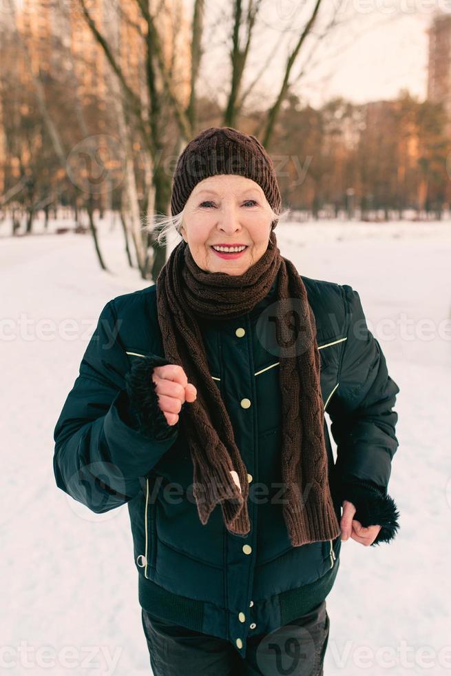 ältere Frau mit Hut und sportlicher Jacke beim Joggen im Schneewinterpark. Winter, Alter, Sport, Aktivität, Saisonkonzept foto