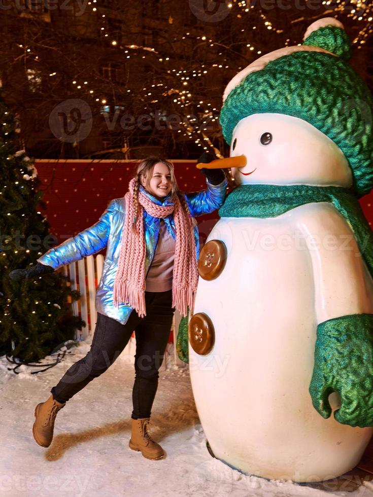 glücklich lächelnde junge kaukasische Frau in Schal, Hut, Jacke, Handschuhe durch den Schneemann im Freien. neues Jahr, Spaß, Winterkonzept foto