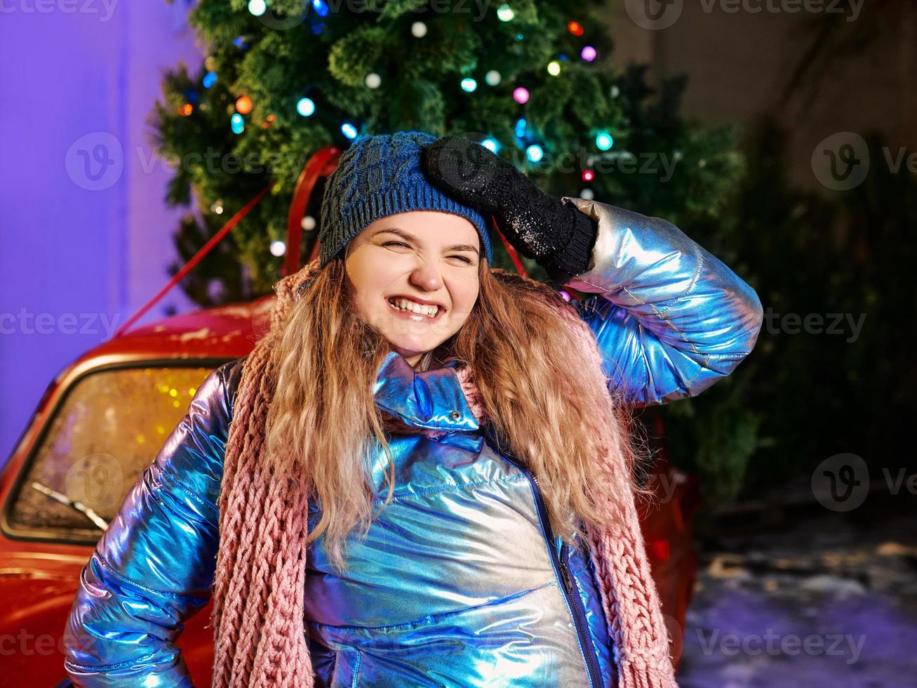 glücklich lächelnde junge kaukasische Frau in Schal, Hut, Jacke, Handschuhe durch den roten Oldtimer und Weihnachtsbaum im Freien. neues Jahr, Spaß, Winterkonzept foto