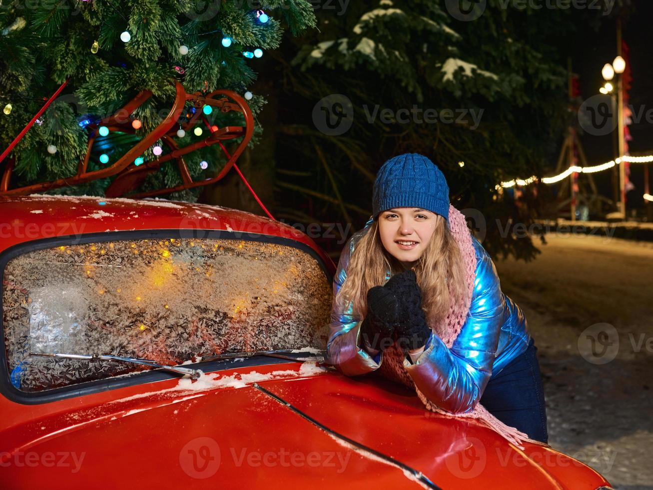 glücklich lächelnde junge kaukasische Frau in Schal, Hut, Jacke, Handschuhe durch den roten Oldtimer und Weihnachtsbaum im Freien. neues Jahr, Spaß, Winterkonzept foto