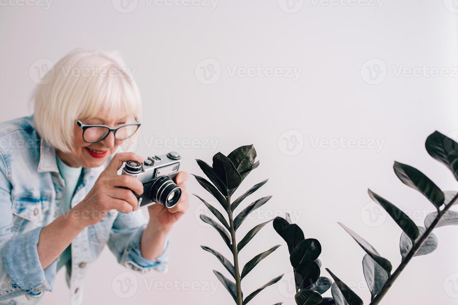 Senior stilvolle Frau mit grauem Haar und in Brille und Jeansjacke, die Blumen mit Filmkamera fotografiert. Alter, Hobby, Anti-Age, positive Stimmung, Fotografiekonzept foto