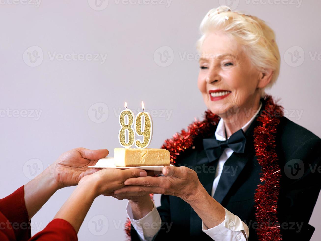 Fröhliche, stilvolle, neunundachtzig Jahre alte Frau im schwarzen Anzug, die ihren Geburtstag mit Kuchen feiert. Lifestyle, Positiv, Mode, Stilkonzept foto