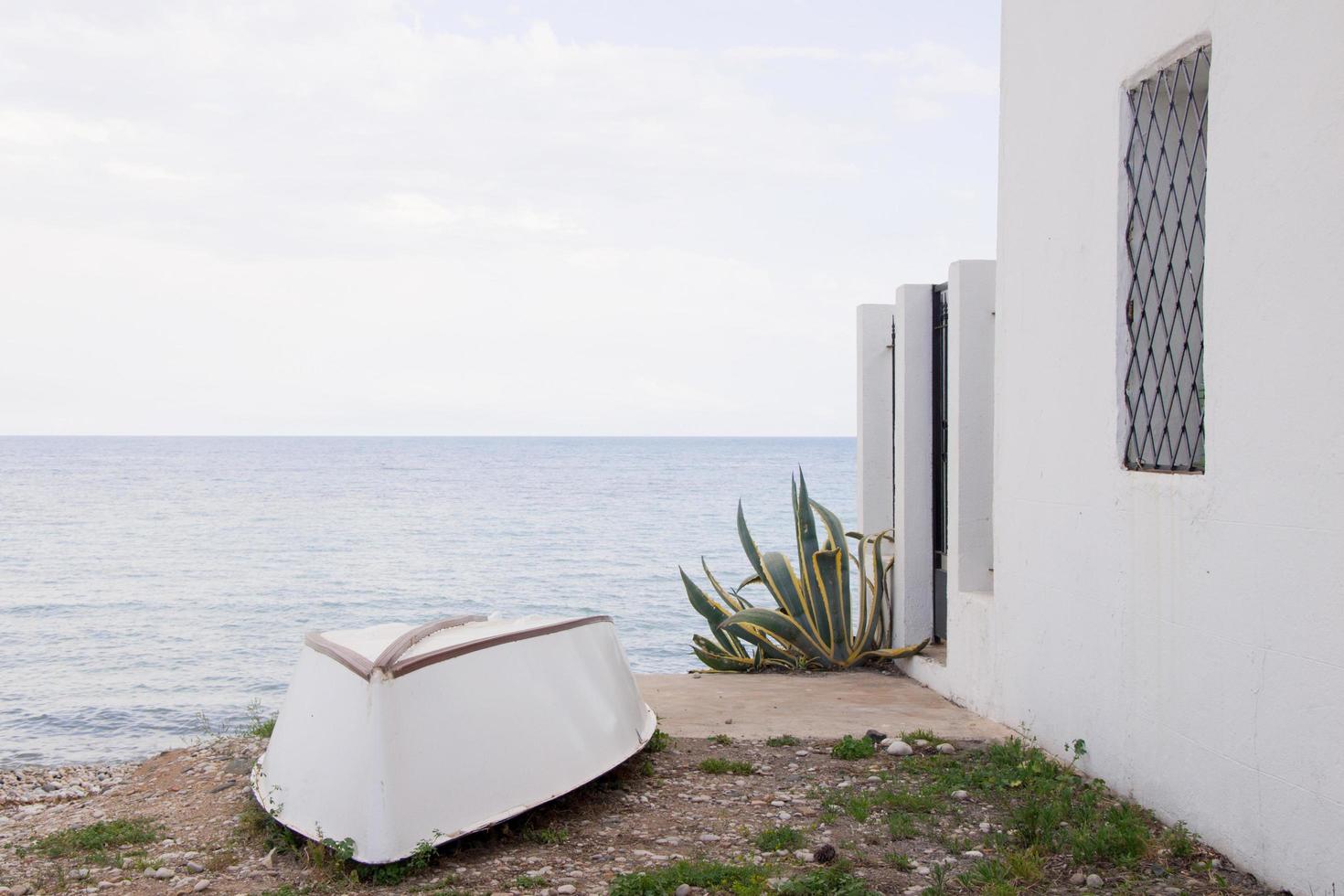 umgedrehtes weißes Boot am Strand, neben einem weißen Haus und einer grünen Pflanze. ruhiges Meer im Hintergrund. altea foto