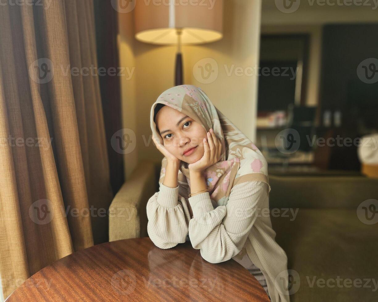 asiatisch Muslim Frau tragen Hijab Sitzung nachdenklich während unterstützen Kinn und Kopf, suchen beim Kamera mit verschwommen Hintergrund foto