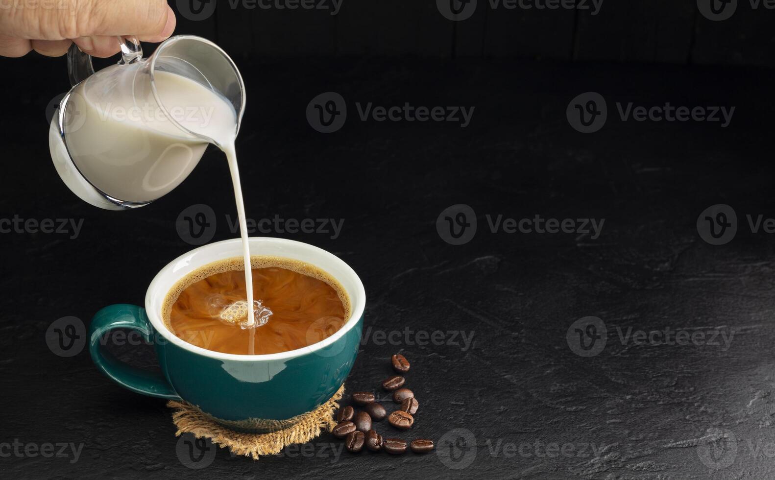 Milch Sahne gegossen in ein Grün Kaffee Tasse auf ein hölzern Tisch, schwarz und dunkel Hintergrund. foto