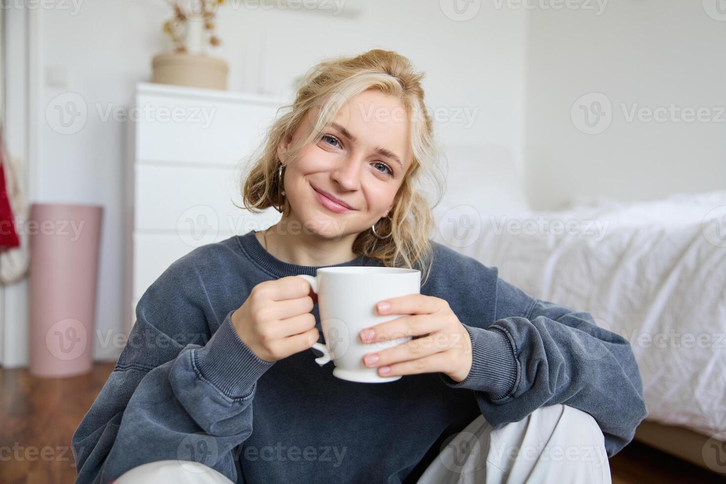Bild von jung Teenager Mädchen Sitzung im ihr Schlafzimmer auf Boden, Trinken Tasse von Tee und genießen Tag beim heim, lächelnd und suchen beim Kamera foto