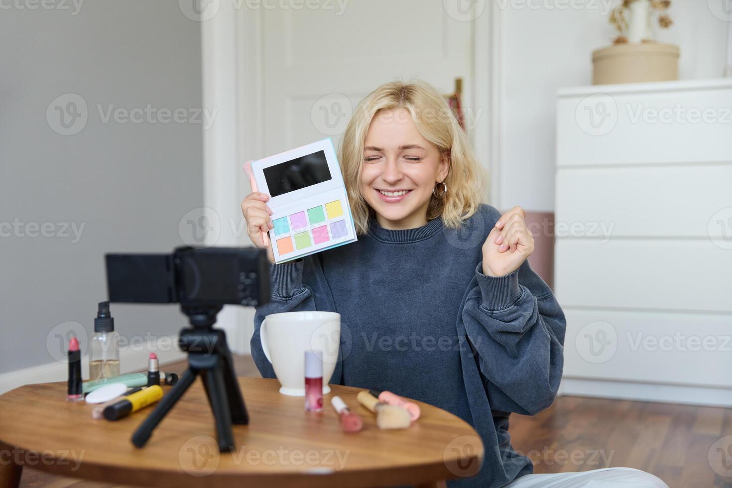 aufgeregt lächelnd blond Frau Aufzeichnung Rezension auf ein Neu Lidschatten Palette, Lebensstil Blogger zeigen Produkt zu Anhänger, sitzt auf Fußboden mit begeistert Gesicht foto