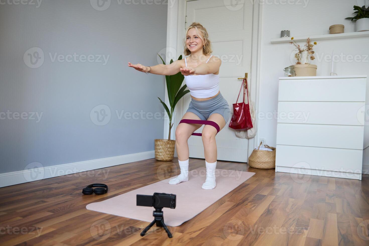 Porträt von Frau, Fitness Lehrer beim heim, Aufzeichnung Über trainieren, zeigen Wie zu tun Bein Übungen, Kniebeugen mit elastisch Widerstand Band, Arbeiten aus drinnen auf Yoga Matte foto