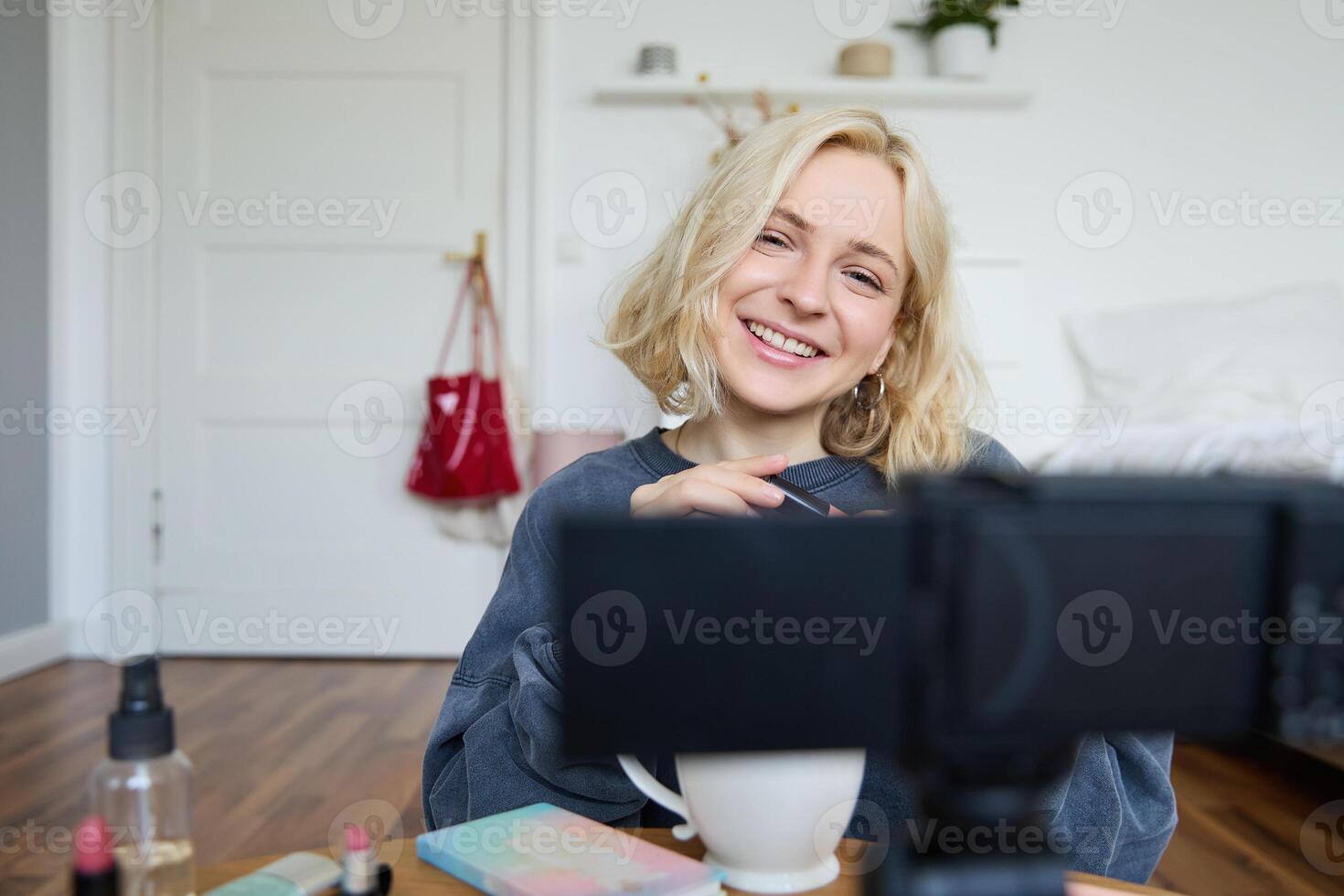 schließen oben Porträt von jung Mädchen Vlogger, Teenager Aufzeichnungen Blog Über bilden, Verwendet Kamera auf Stabilisator, Sitzung auf Fußboden in der Nähe von Kaffee Tisch, bewirbt sich Wimperntusche und kosmetisch Produkte foto