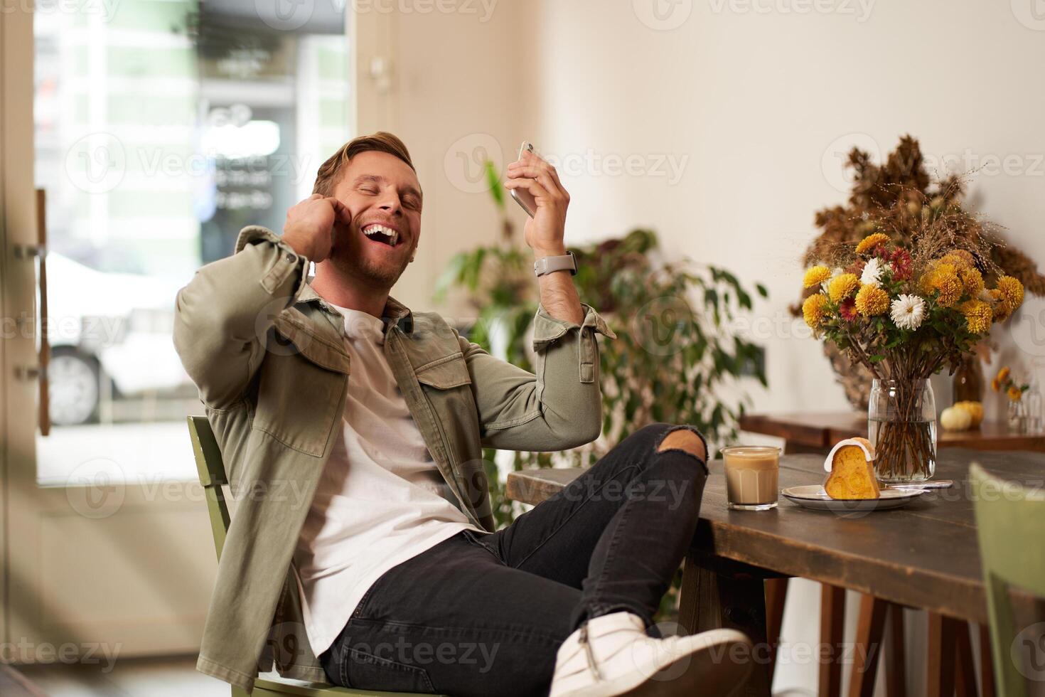 Lebensstil Porträt von gut aussehend jung Mann, Sitzung im Cafe, reden zu ein Freund auf Handy, Mobiltelefon Telefon mit kabellos Kopfhörer, Lachen und lächelnd, Hören zu komisch Podcast foto