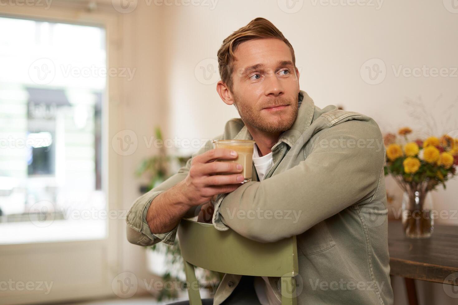 Porträt von gut aussehend jung Mann mit Tasse von Kaffee, Sitzung auf Stuhl im Cafe, lächelnd und entspannend mit seine Cappuccino trinken foto