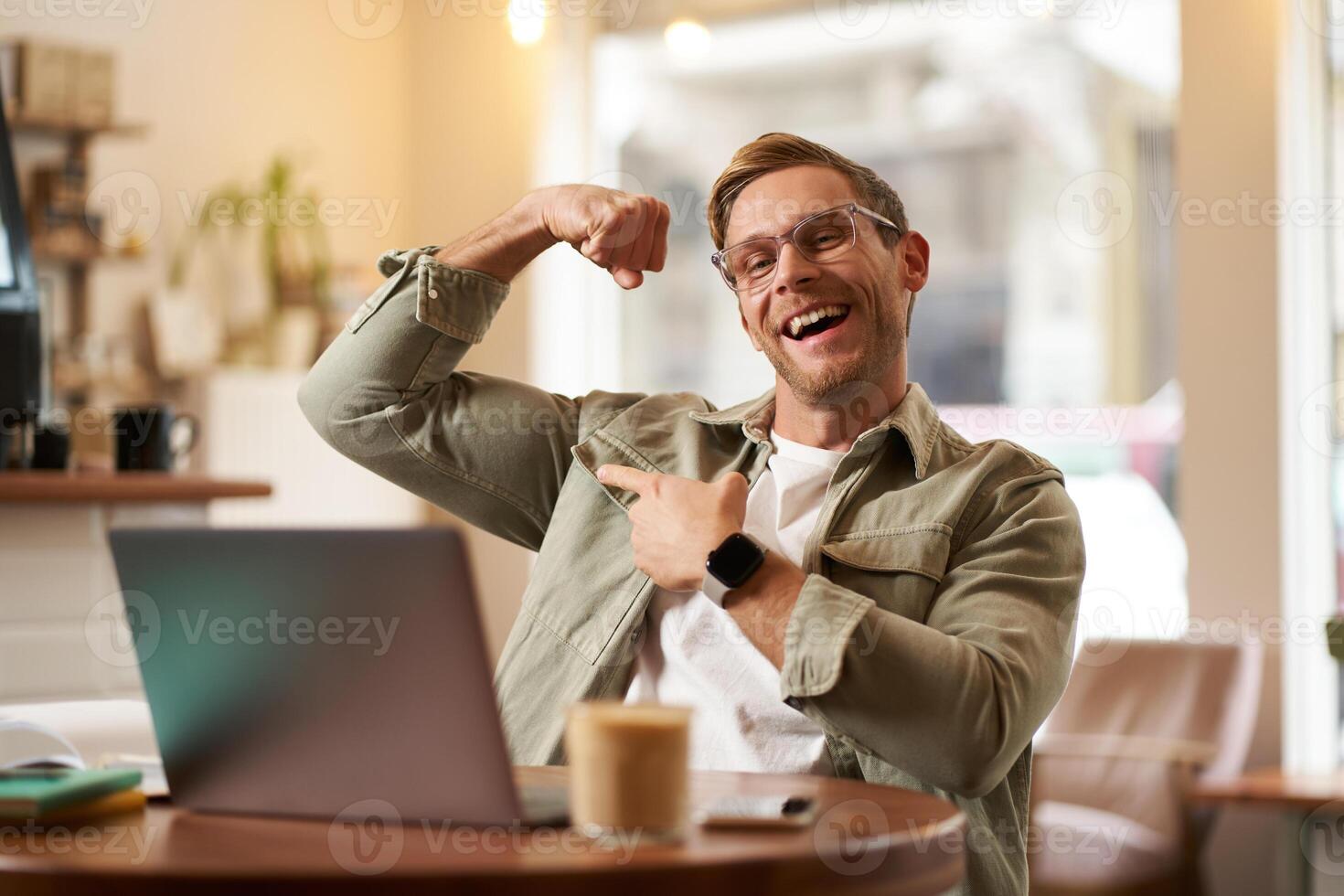 Porträt von gutaussehend, zufrieden jung Mann zeigt an Bizeps, Biegen Muskeln, sitzt im Vorderseite von Laptop im Cafe, Jubel, Privat Trainer haben ein Plaudern mit seine Klient foto