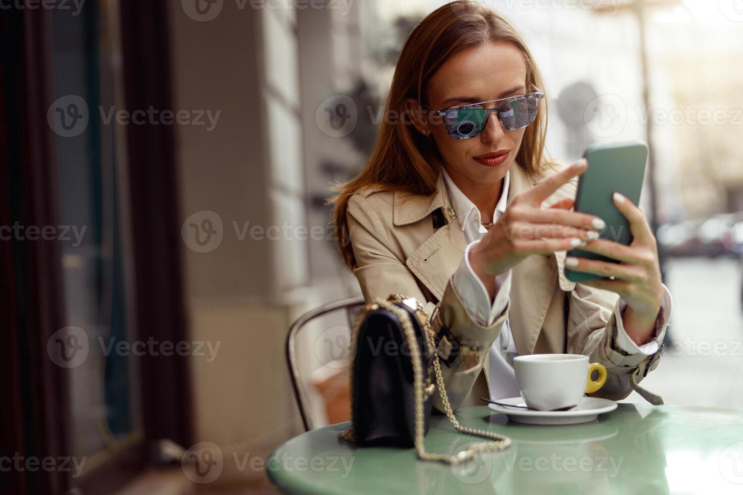 lächelnd stilvoll Frau im Brille Sitzung beim Cafe Terrasse und halten Telefon während Trinken Kaffee foto