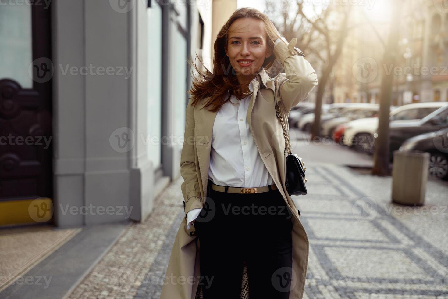 attraktiv Frau im Mantel Kreuzung das Stadt Straße und suchen beim Kamera. verschwommen Hintergrund foto