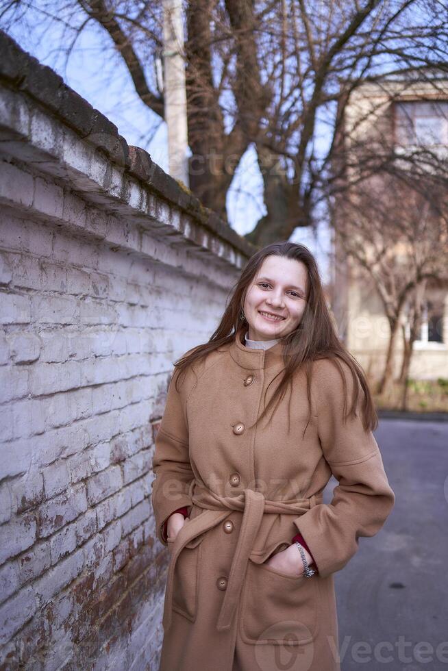 ein Mädchen im ein braun Mantel ist glücklich in der Nähe von ein Weiß Mauer auf ein Frühling Tag foto