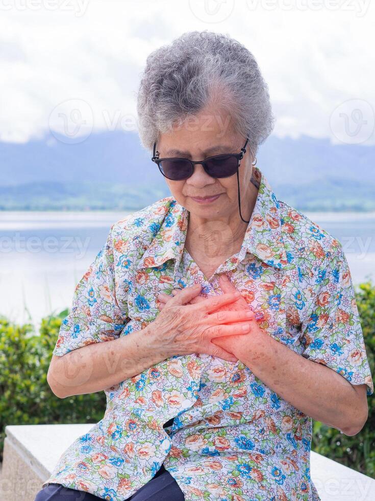 ein Senior Frau umklammern ihr Truhe im Schmerzen beim das Zeichen von Angina oder Myokard Infarkt oder Herz Attacke während Sitzung beim das Seite von das See. Konzept von alt Menschen und Herz Attacke foto