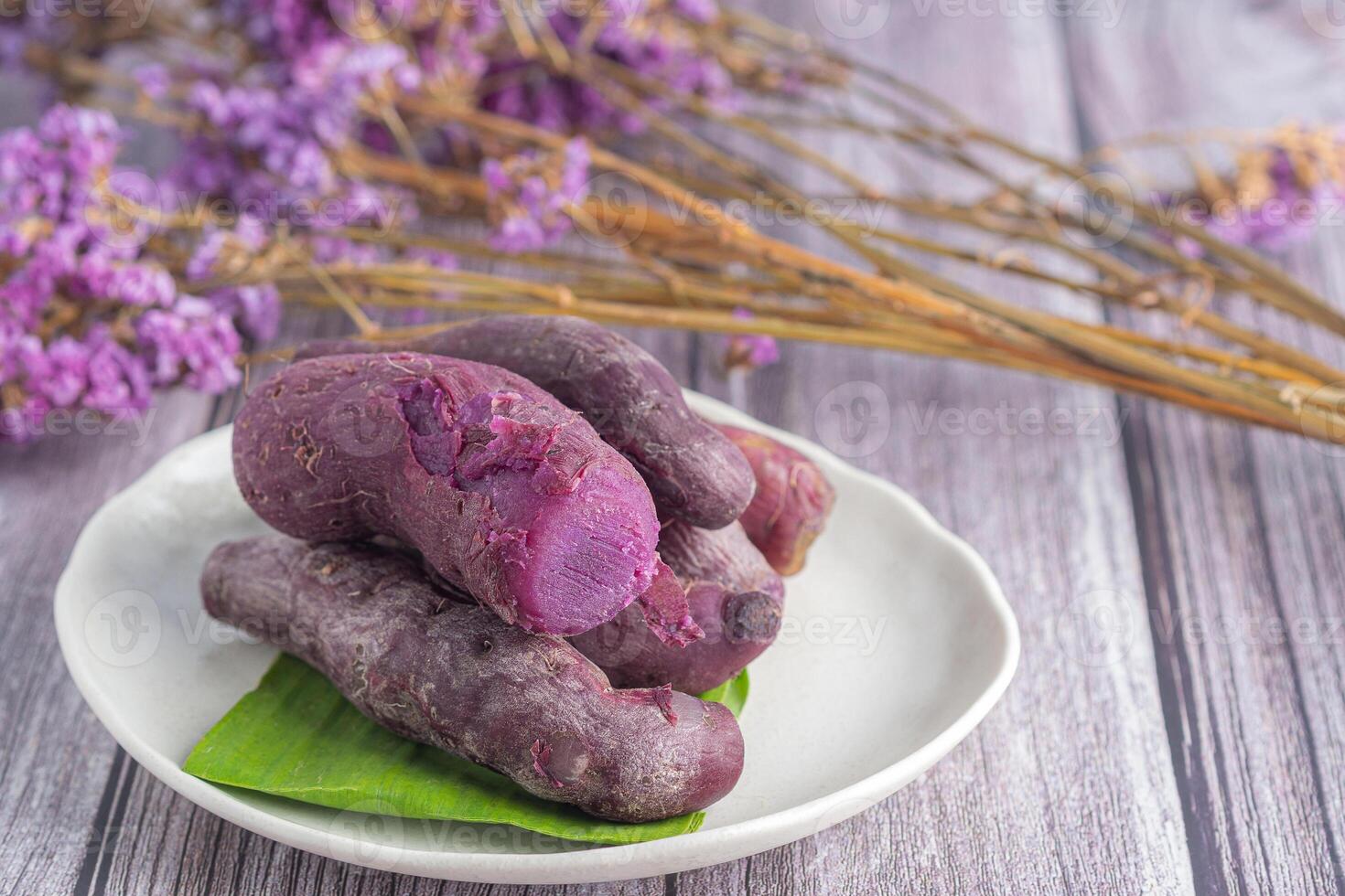 ein Stapel von reif lila Süßkartoffeln ist platziert auf ein Teller auf ein hölzern Tabelle mit verschwommen lila Blumen im das Hintergrund. Raum zum Text. Konzept von Gesundheit Obst foto