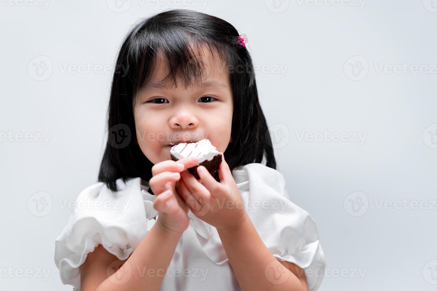 glückliches Mädchen beißt süßen Schokoladenkuchen. sie trägt weißes hemd. süßes Lächeln. Kinderkonzept mit Süßigkeiten essen. foto