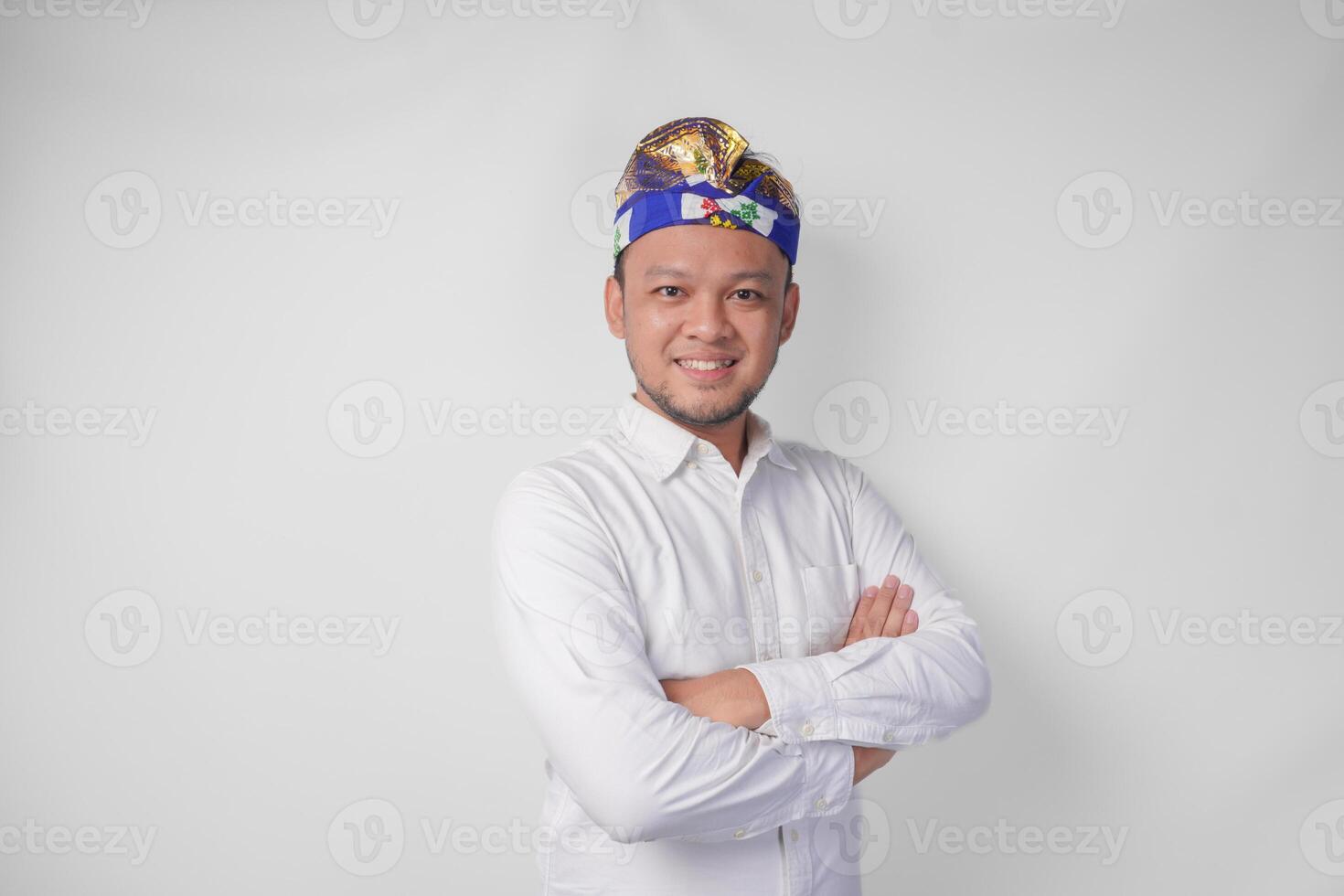 Porträt von ein balinesisch tragen traditionell Kopfschmuck namens udeng posieren mit ein gekreuzt Waffen und zuversichtlich Lächeln Über isoliert Weiß Hintergrund foto