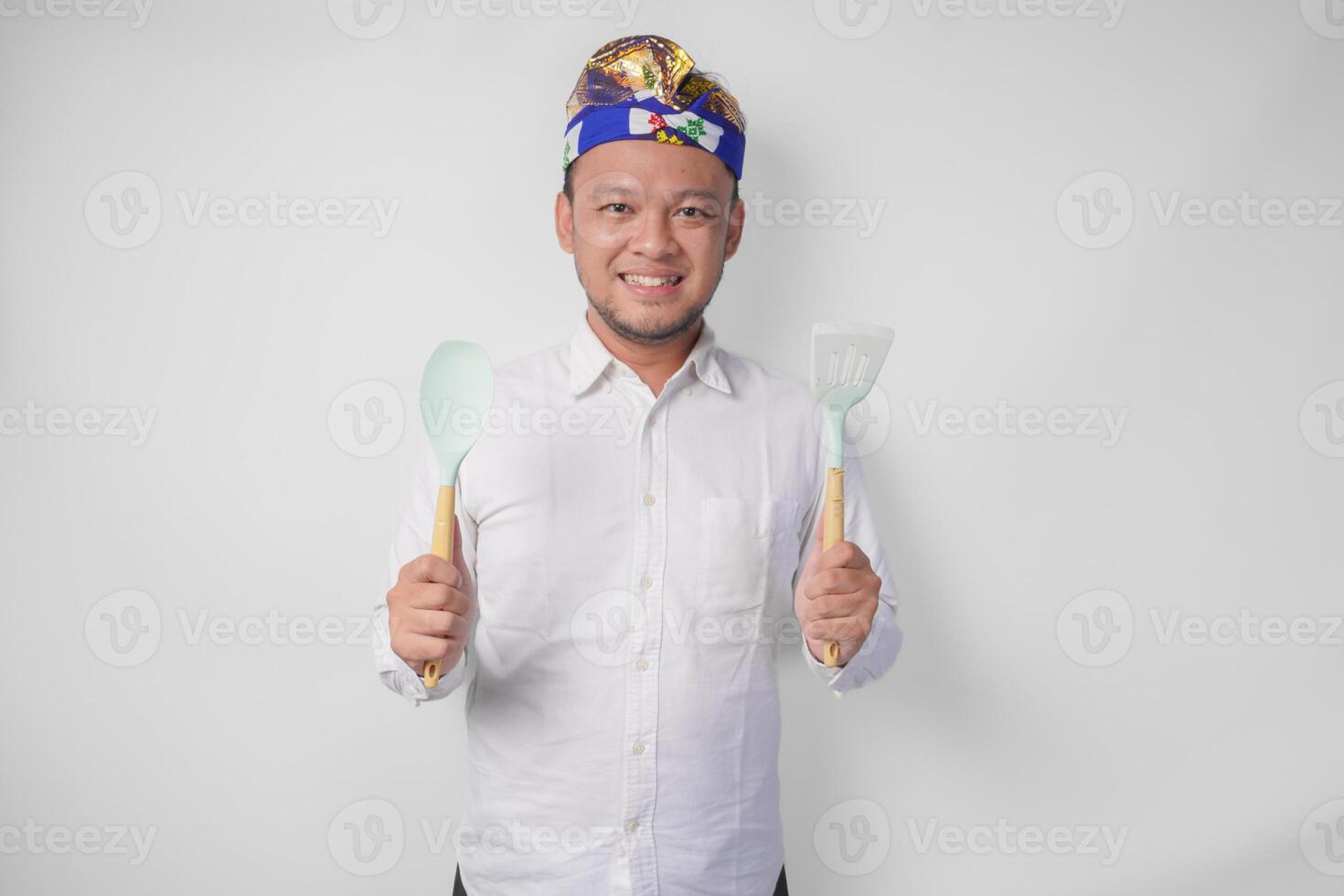 lächelnd jung balinesisch Mann im Weiß Hemd und traditionell Kopfschmuck halten Spatel und Küche Kochen Utensilien foto