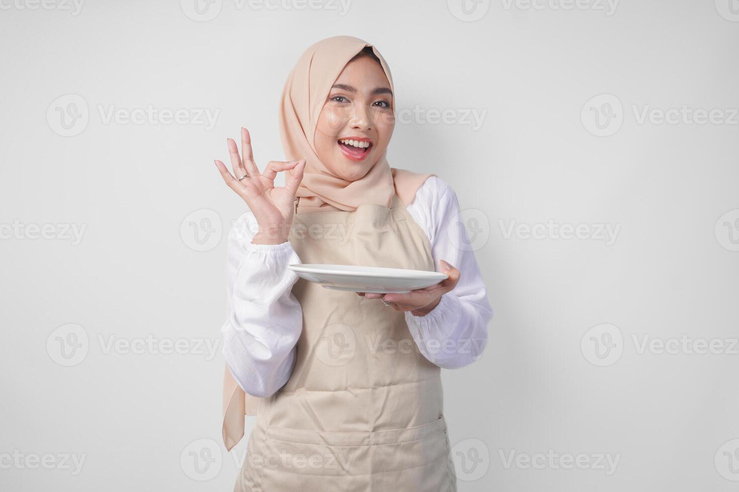 glücklich jung asiatisch Muslim Frau im Hijab und Sahne Schürze halten ein leeren Teller mit Kopieren Raum während Herstellung ein köstlich Hand Geste zu ausdrücken Wie gut das Essen ist foto
