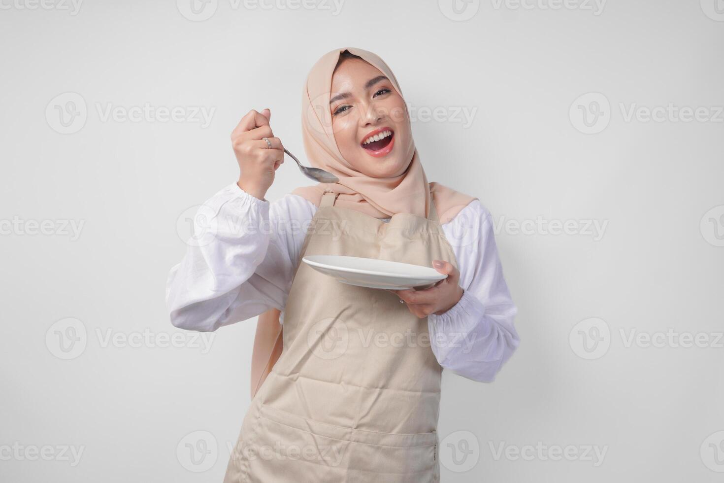 hungrig jung asiatisch Muslim Frau im Hijab und Sahne Schürze halten Löffel und Essen von ein leeren Teller mit Kopieren Raum Über Es. Ramadan Konzept foto