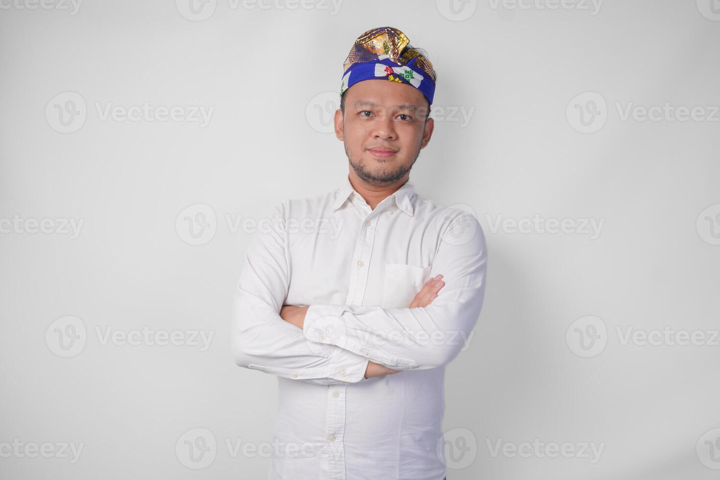 Porträt von ein balinesisch tragen traditionell Kopfschmuck namens udeng posieren mit ein gekreuzt Waffen und zuversichtlich Lächeln Über isoliert Weiß Hintergrund foto