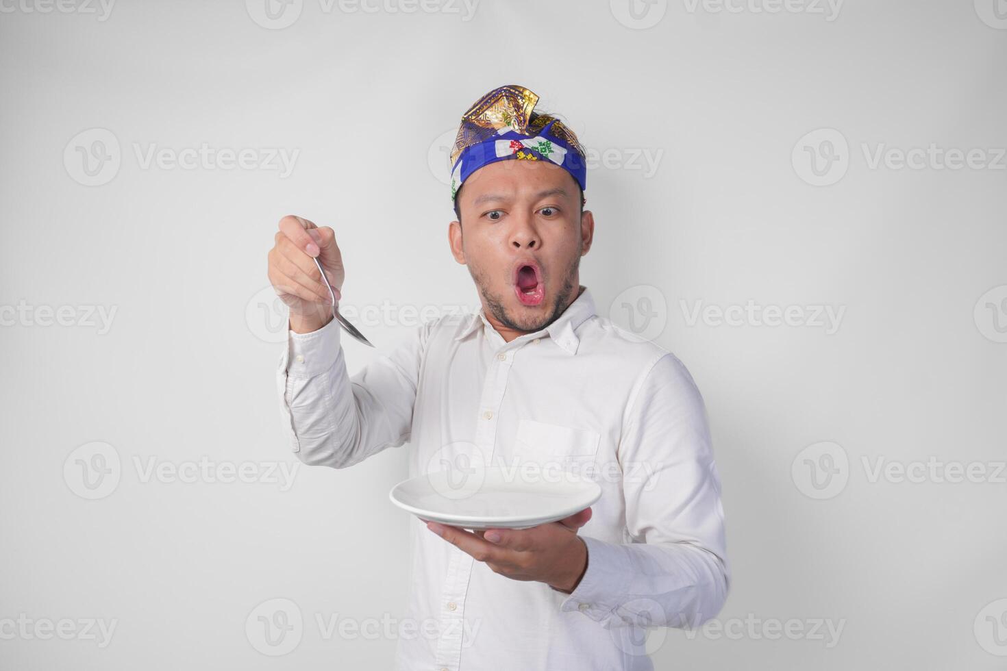 schockiert jung balinesisch Mann im Weiß Hemd und traditionell Kopfschmuck halten ein leeren Teller mit Kopieren Raum und Löffel, Gefühl überrascht durch das Essen Speisekarte foto