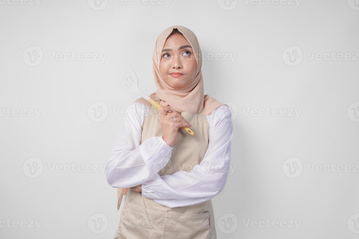 nachdenklich jung asiatisch Muslim Frau im Hijab und Sahne Schürze halten Spatel und Küche Kochen Utensilien, Denken schwer Was Essen Speisekarte zu Koch foto