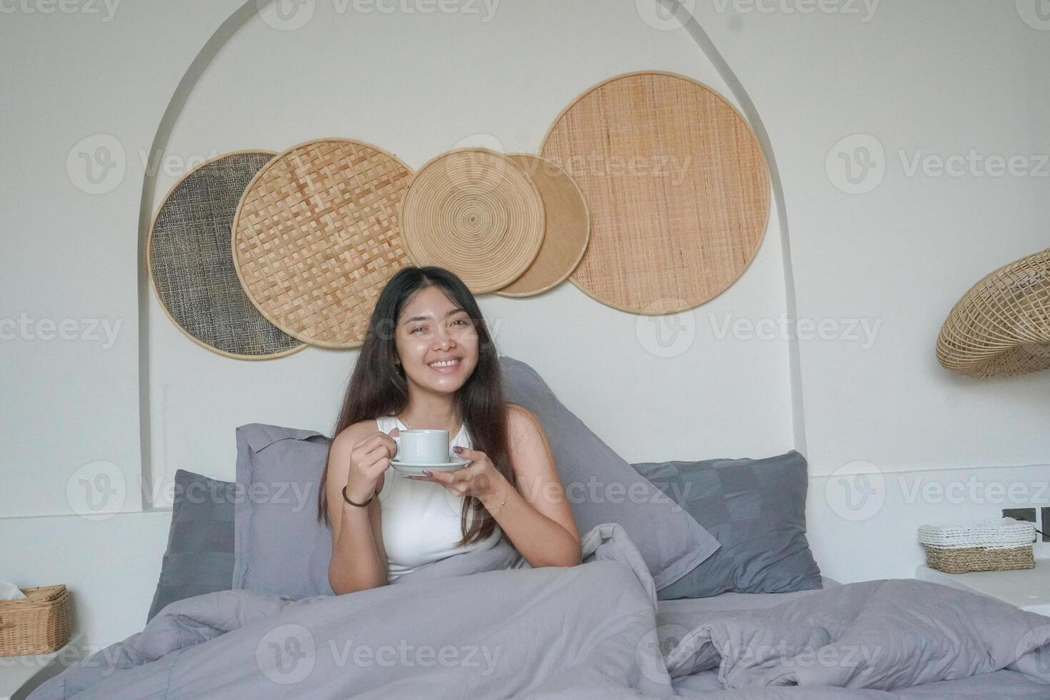 glücklich attraktiv asiatisch Frau im Weiß oben posieren beim das Schlafzimmer nach aufwachen hoch, lächelnd fröhlich während halten Tasse von Tee. Urlaub Freizeit Konzept. foto