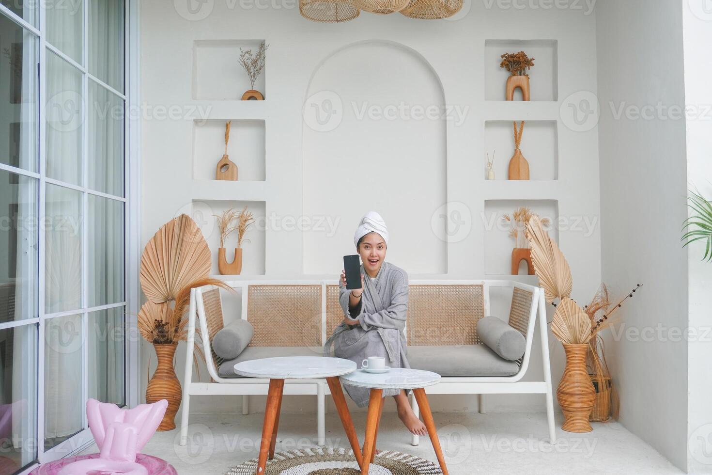 glücklich asiatisch Frau tragen grau Bademantel Sitzung auf ein Couch, zeigen Clever Telefon Bildschirm während lächelnd fröhlich. Urlaub Freizeit Konzept. foto