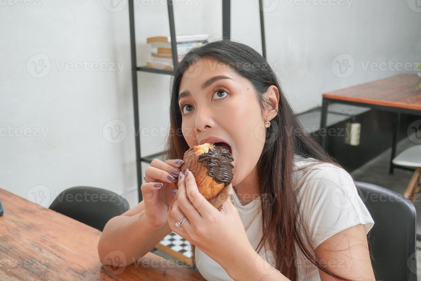 schön asiatisch jung Frau Essen köstlich Gebäck genannt Cromboloni beim ein Café. das Dame beißt Stück von Schokolade Füllung Cromboloni, suchen glücklich beim das Cafe foto