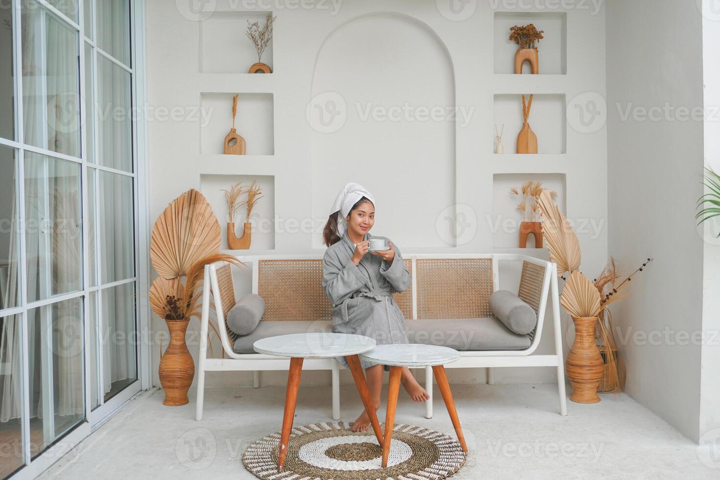 glücklich attraktiv asiatisch Frau im grau Bademantel posieren beim das Terrasse, lächelnd beim Kamera während Trinken Tee. Urlaub Freizeit Konzept. foto