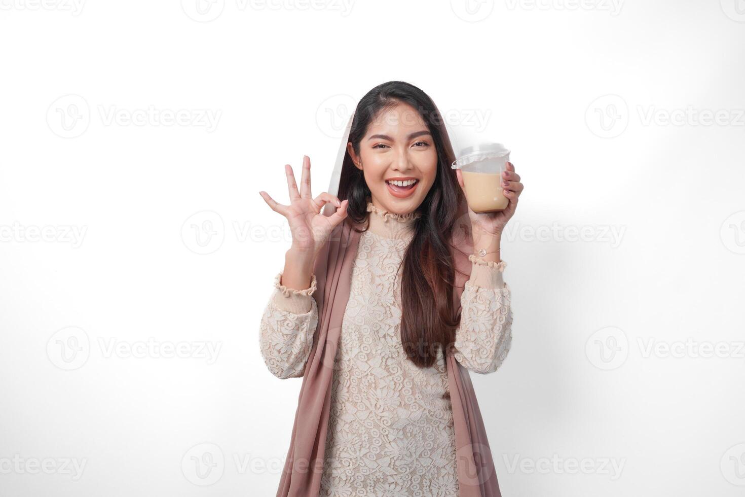 heiter asiatisch Muslim Frau im Kopftuch Schleier Hijab halten Kaffee im ein Plastik Tasse warten zum iftar zu trinken während Herstellung ein okay Zeichen Geste. Ramadan Konzept foto