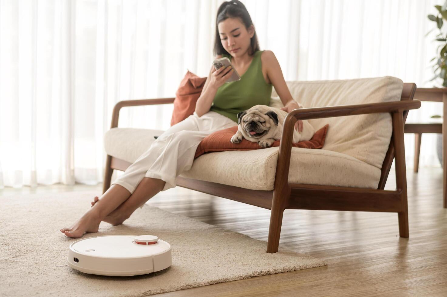 jung asiatisch Frau entspannend mit Mops Hund im Leben Zimmer während Roboter Vakuum Reiniger Arbeiten foto