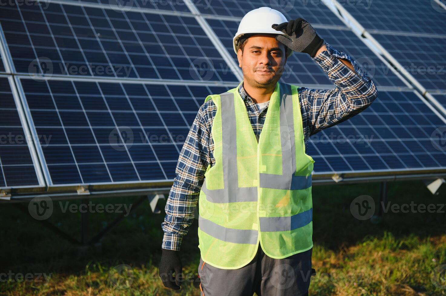 Porträt von jung indisch Mann Techniker tragen Weiß schwer Hut Stehen in der Nähe von Solar- Paneele gegen Blau Himmel. industriell Arbeiter Solar- System Installation foto