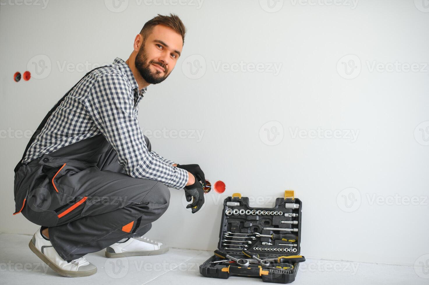 Elektriker Baumeister beim arbeiten, Installation von Steckdosen und Schalter. Fachmann im Overall mit ein Elektriker Werkzeug. gegen das Hintergrund von das Reparatur Seite? ˅ foto