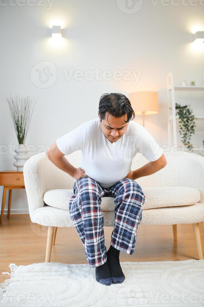 unzufrieden indisch oder arabisch Mann, sitzt auf komfortabel Sofa im gemütlich Leben Zimmer, hält seine Hände auf seine Magen, Grimassen von Schmerzen im seine Magen, leidet von Vergiftung, Krampf, Bauch Probleme. foto