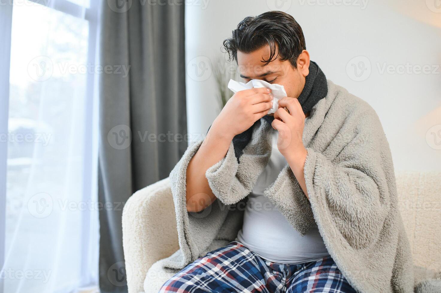 krank Mann Sitzung beim Zuhause auf Sofa im Leben Zimmer, Niesen hat Allergie und flüssig Nase, indisch Mann mit Serviette hat ein kalt foto