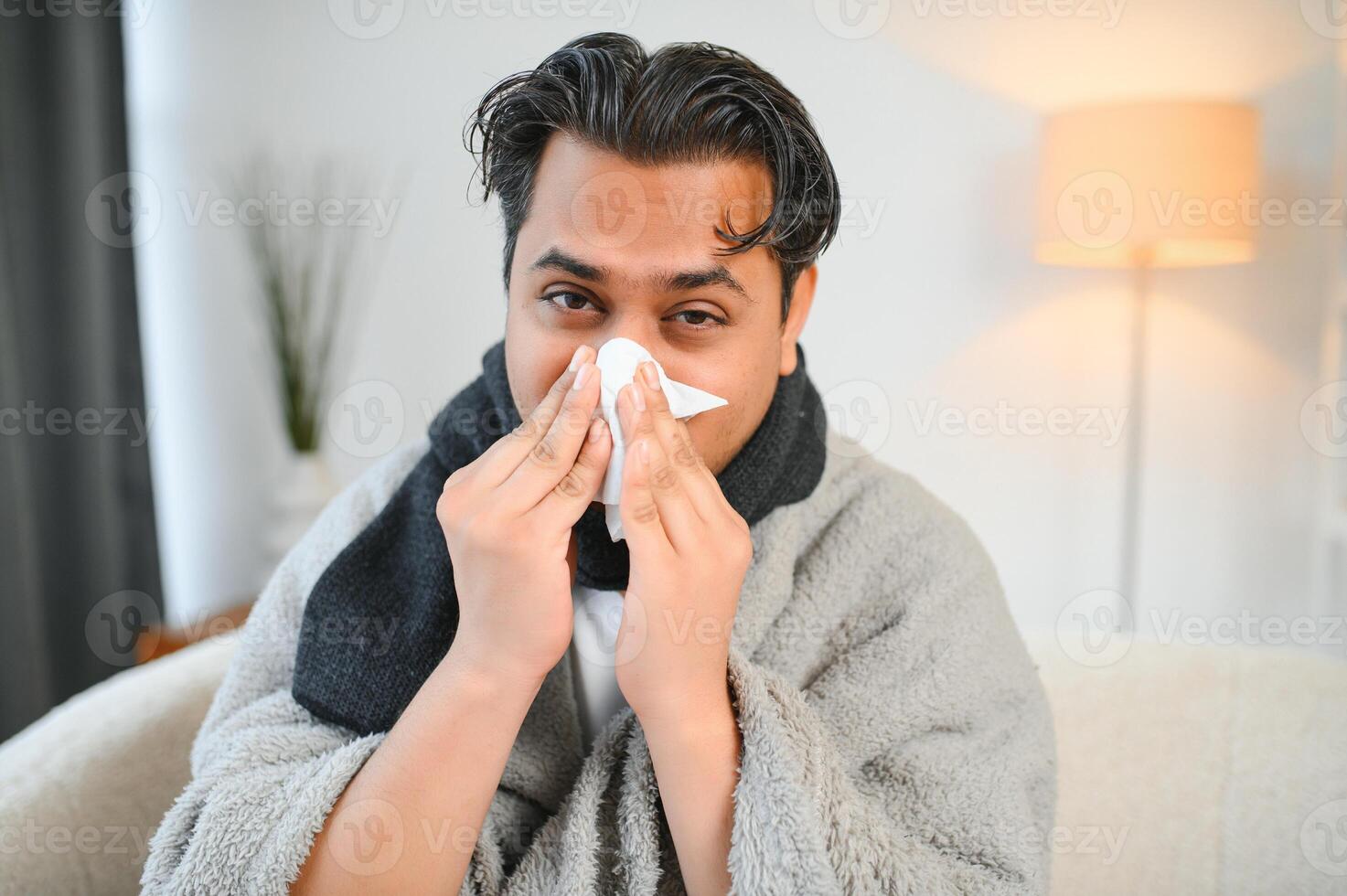 krank Mann Sitzung beim Zuhause auf Sofa im Leben Zimmer, Niesen hat Allergie und flüssig Nase, indisch Mann mit Serviette hat ein kalt foto
