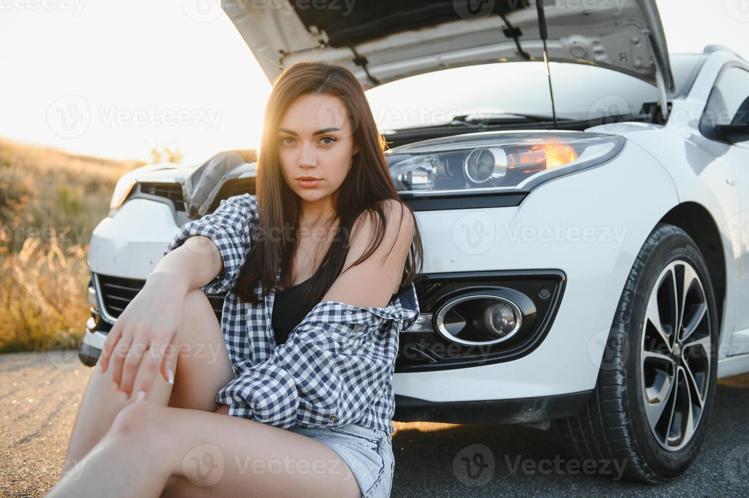 ein jung Mädchen sitzt in der Nähe von ein gebrochen Auto auf das Straße mit ein öffnen Haube. foto