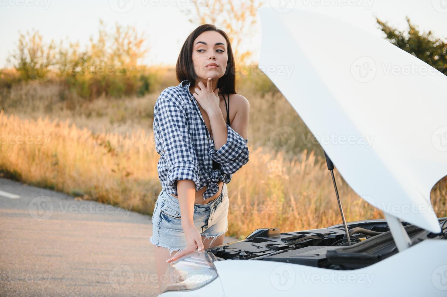schön schlank Mädchen im Hemd und kurze Hose sieht aus im öffnen Auto Kapuze auf ein Straße foto