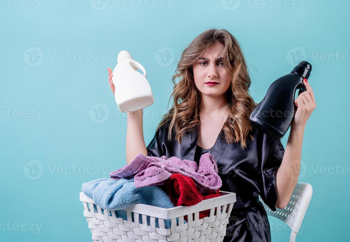 verwirrte Hausfrau, die einen Korb mit Kleidung und Waschmittelflaschen hält, die auf blauem Hintergrund zum Waschen bereit sind foto