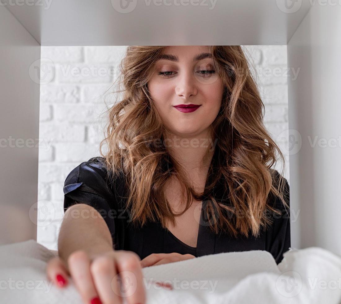 lächelnde junge Frau, die einen Stapel Handtücher auf einem Regal auf weißem Ziegelsteinhintergrund anordnet foto